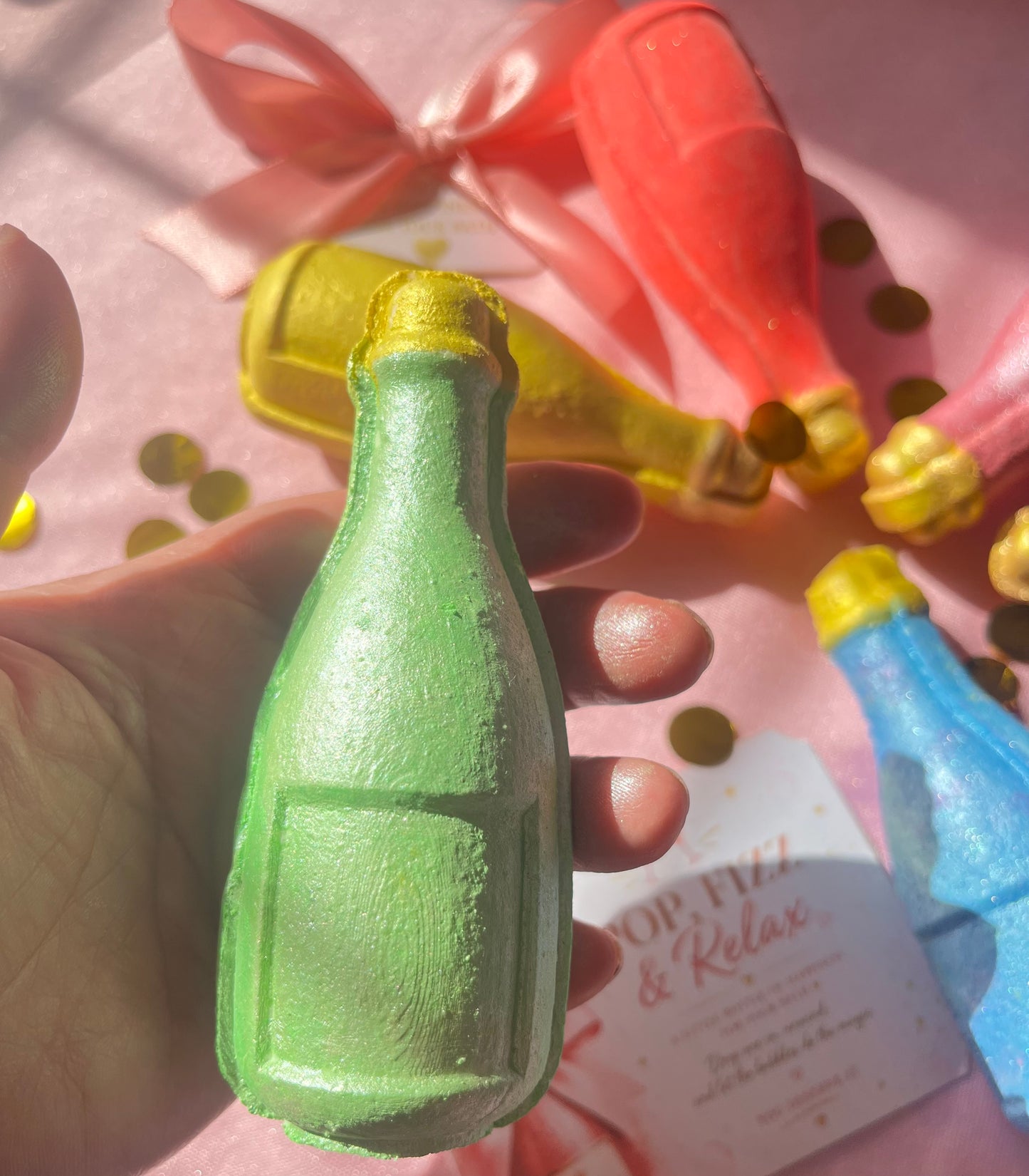 Green bottle-shaped bath bomb held in a hand with colorful bath bombs and confetti in the background.