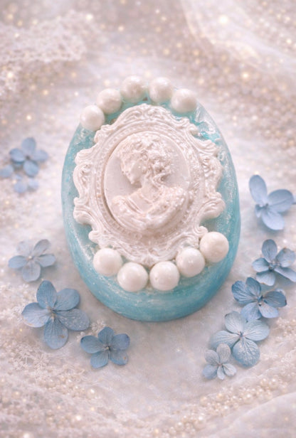 Decorative soap with a vintage-style face design and pearl embellishments, surrounded by small blue flowers on a textured white background.