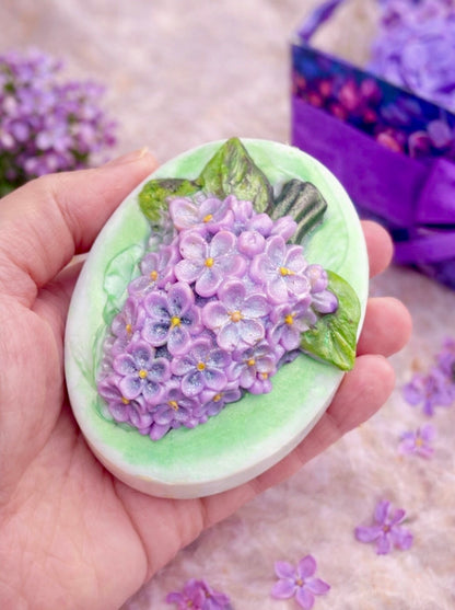 Handmade lilac flower soap held in hand to show size, featuring realistic purple blossoms and green leaves, part of the Lilac Spa Gift Box for her.

