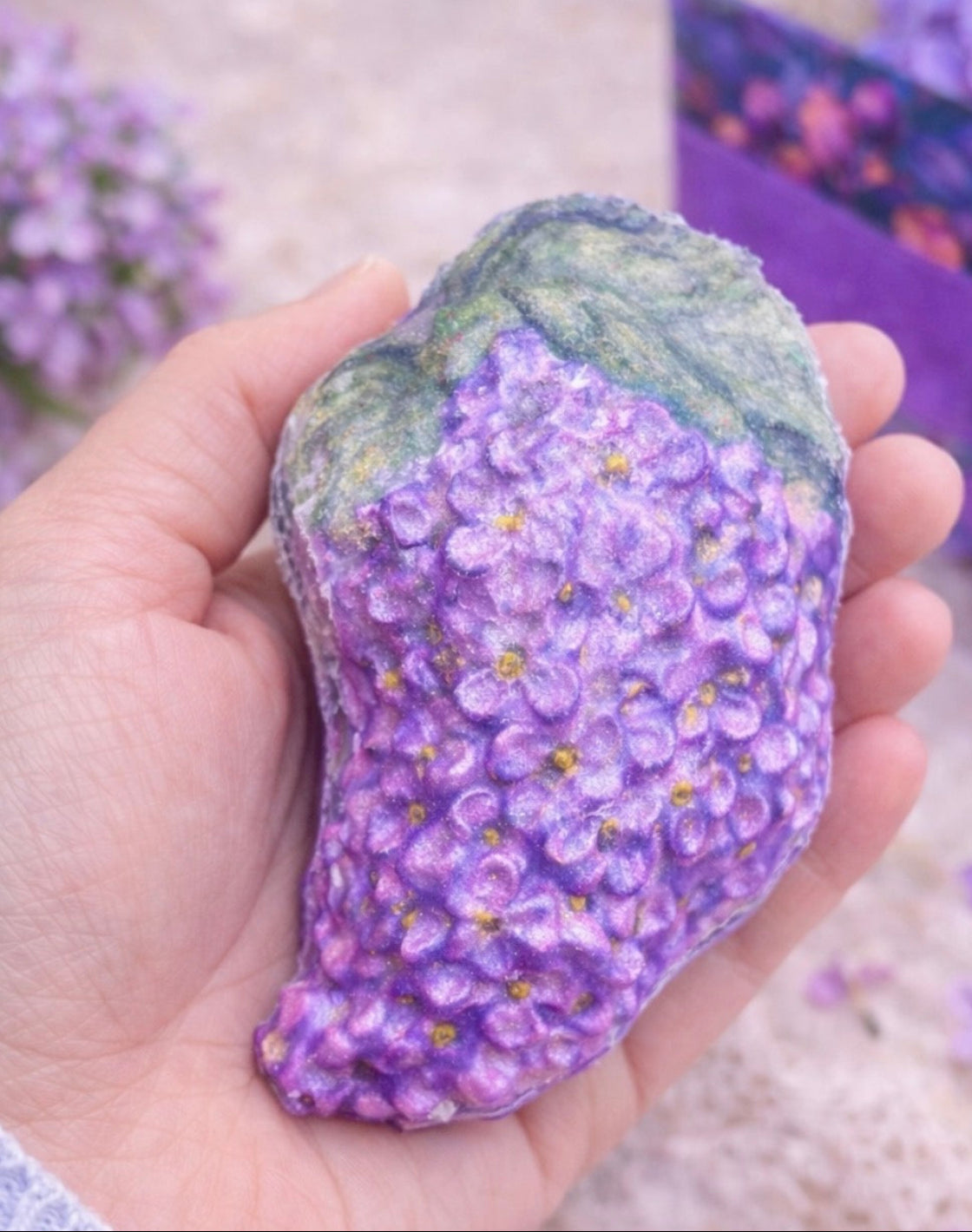 Lilac bath bomb held in hand to show size, featuring realistic purple lilac blossoms and green leaves, handmade spa treat from the Lilac Spa Gift Box.