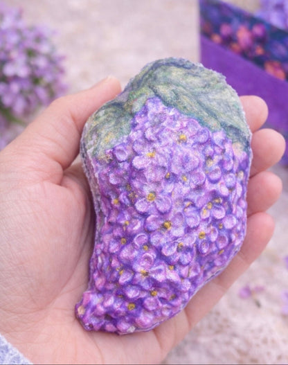 Lilac bath bomb held in hand to show size, featuring realistic purple lilac blossoms and green leaves, handmade spa treat from the Lilac Spa Gift Box.