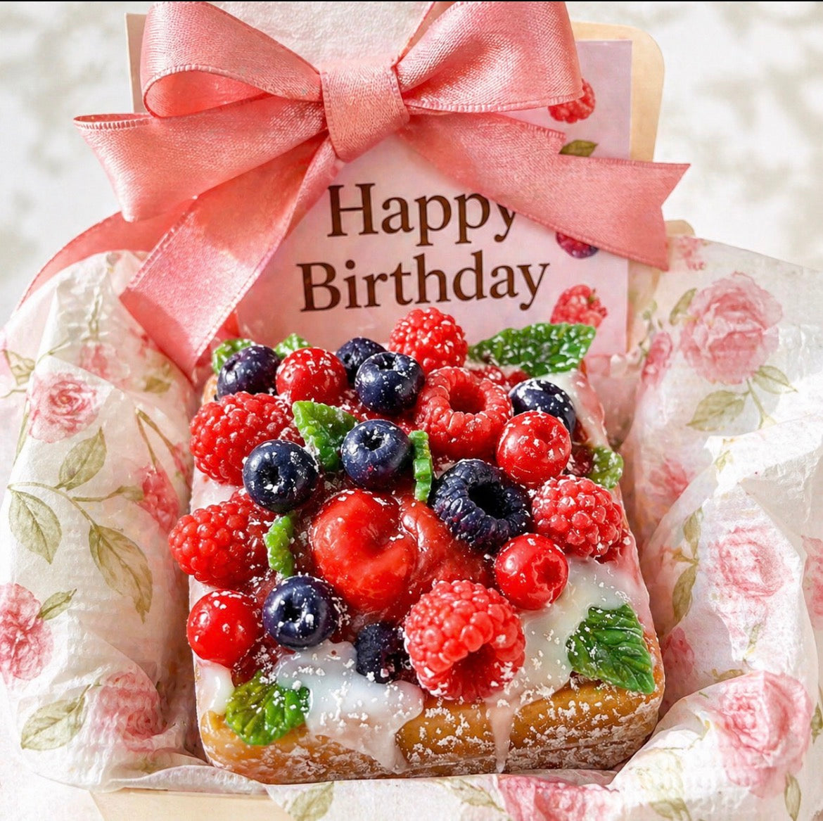 A gift box containing dessert soap with berries on top and a 'Happy Birthday' card, wrapped in floral paper.