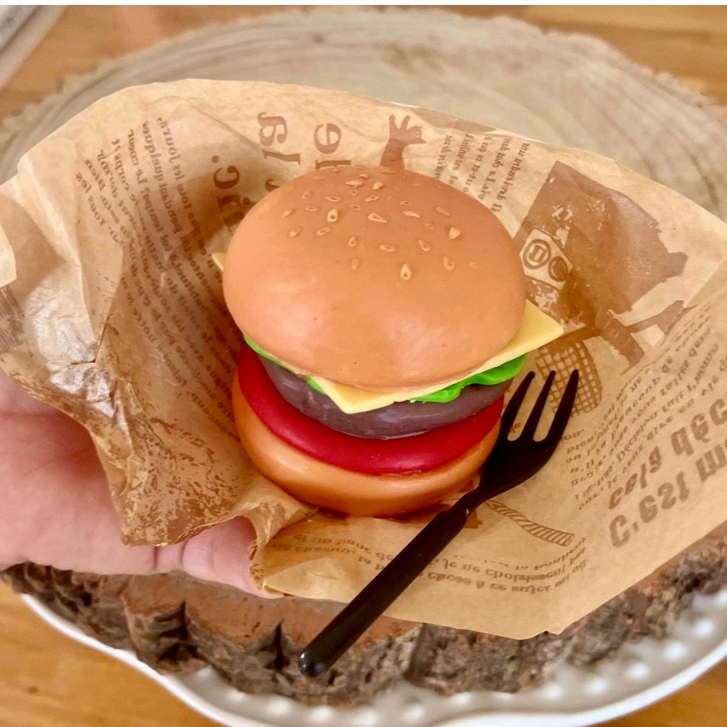 Hand holding a realistic handmade cheeseburger soap with burger layers and sesame bun. Unique novelty soap designed to look like real fast food.