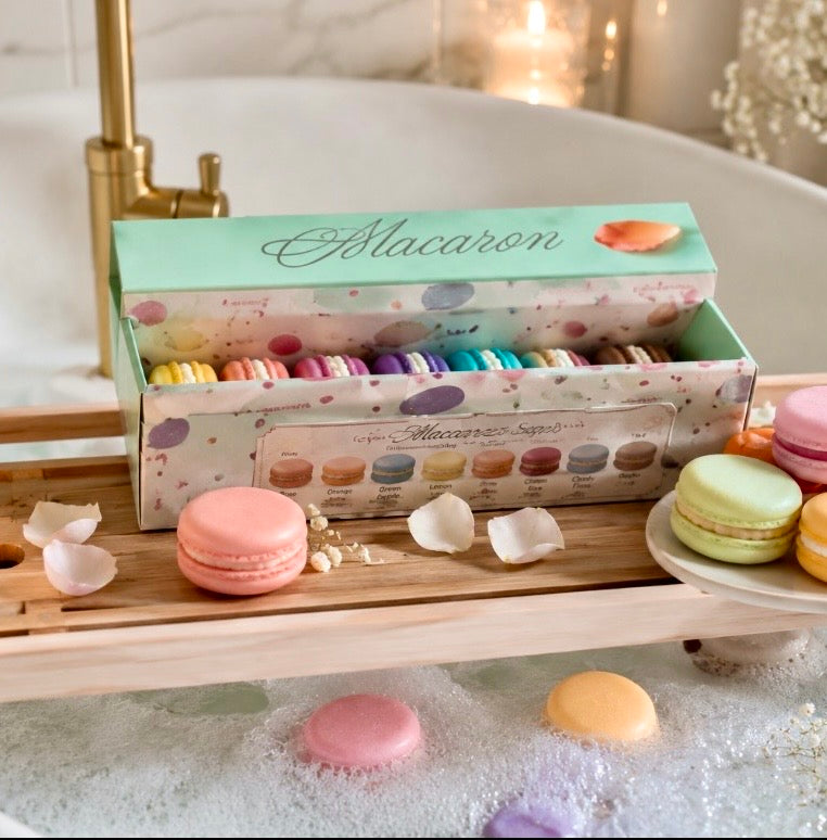 Colourful macaron-shaped soaps displayed with their gift box on a wooden bath tray beside a bubble-filled bathtub.