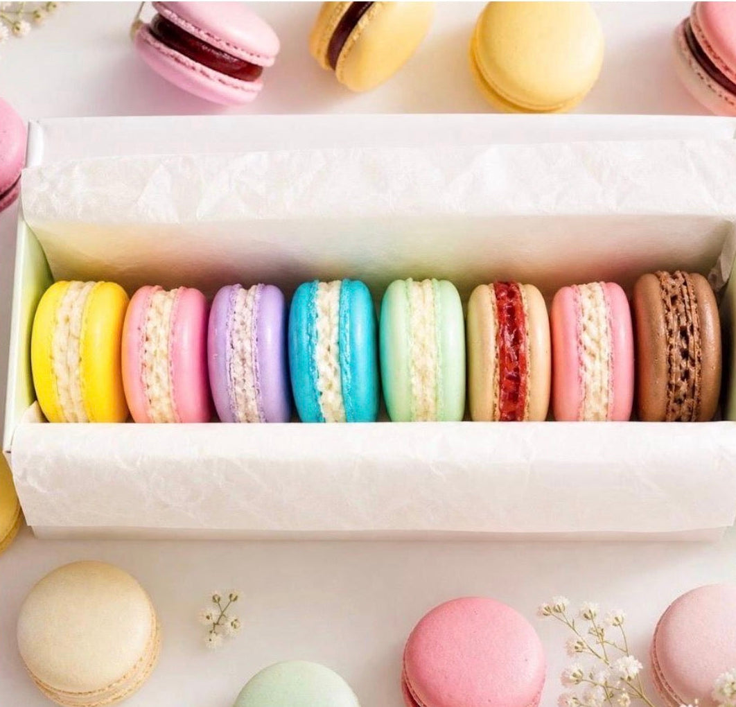 Eight colourful handmade macaron-shaped soaps arranged in a white gift box, surrounded by pastel macaron soaps and delicate flowers.