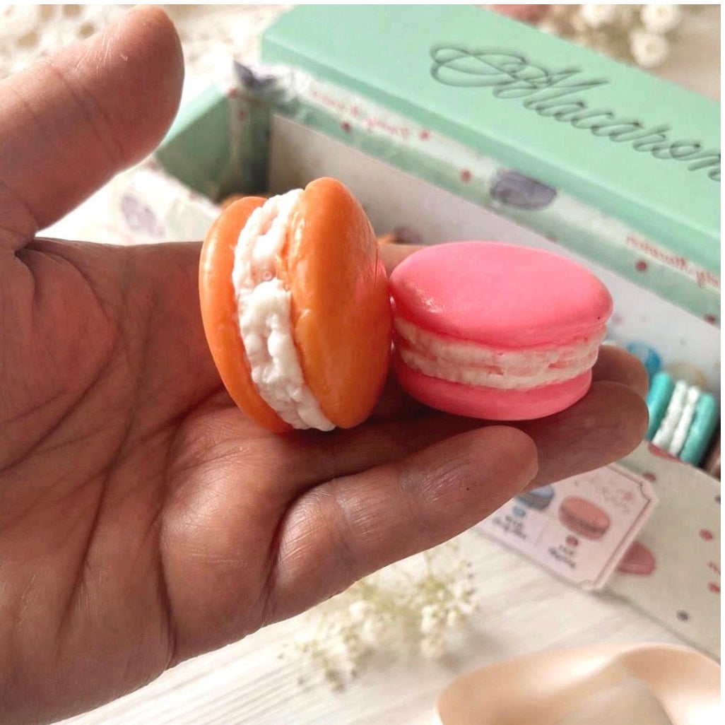 Orange and pink handmade macaron-shaped soaps held in a hand, showing their realistic size and creamy filling details.