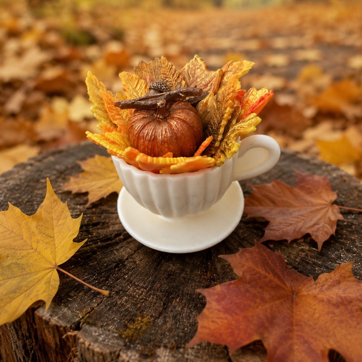 Handmade autumn teacup soap featuring a pumpkin and colourful fall leaves, displayed on a rustic tree stump surrounded by golden autumn leaves. Cozy seasonal gift and autumn home décor.