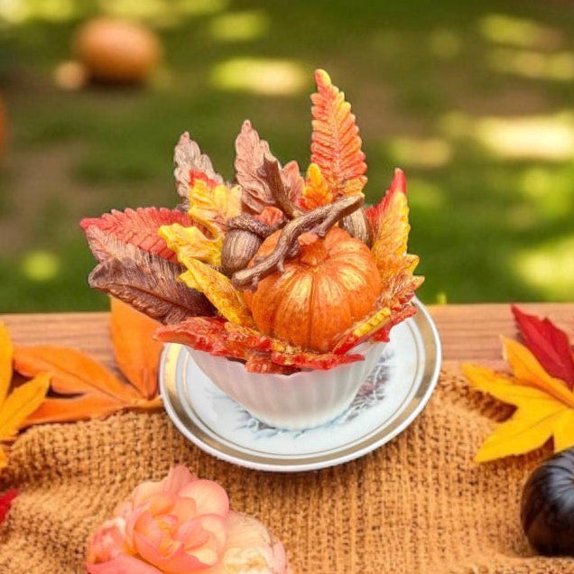 Handmade autumn teacup soap with a pumpkin, colourful fall leaves and woodland details, displayed on a rustic wooden table surrounded by vibrant autumn foliage. Charming seasonal gift for autumn lovers.