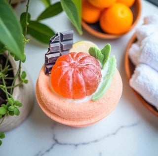 Dessert-style bath bomb with mandarin and chocolate decoration surrounded by oranges and a plant. 