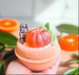 Handmade bath bomb with a mandarin soap on top held by a hand, surrounded by more mandarins and leaves.
