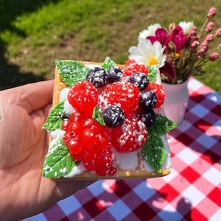 Decorative square waffle soap with berries held by a hand on a checkered tablecloth.