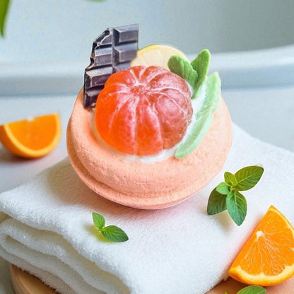 Handmade bath bomb shaped like a dessert with a mandarin, chocolate piece and mint leaves on top, placed o a soft towel with orange slices, spa aesthetic.