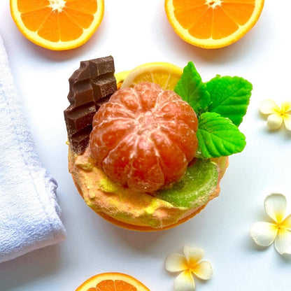 Handmade bath bomb decorated with an mandarin, chocolate, and mint leaves on a white background with flowers.